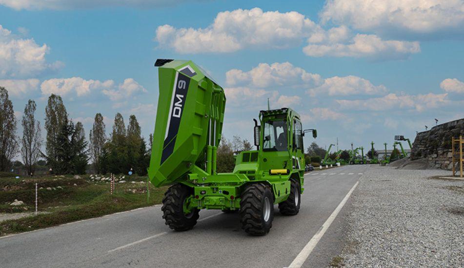 Una macchina dumper MERLO DM9 verde con il cassone sollevato, ferma su una strada circondata da alberi e un cielo nuvoloso sullo sfondo.