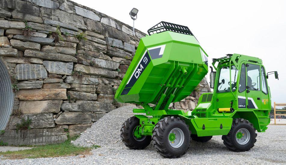 Una macchina dumper MERLO DM9 verde con il cassone sollevato, ferma su ghiaia accanto a un muro di pietra.